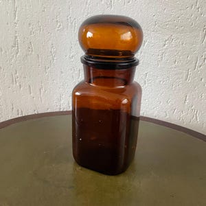 Op de afbeelding: Een vintage apothekerspot van amberkleurig glas met een afgeronde stop. De pot heeft een vierkante basis en een cilindrisch lichaam. De stop is een donkerdere tint amber. De pot staat op een ronde tafel.