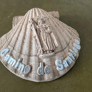 May include: A white scallop shell with a raised image of a pilgrim and the text "Camino de Santiago" in silver lettering.
