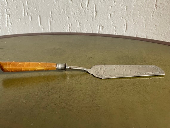 Schmaler Tortenheber mit bernsteinfarbenem Griff – sehr fein mit symmetrischen Motiven graviert. 21 cm. Um 1930