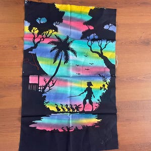 Op de afbeelding: Zwarte stof met een kleurrijke regenbooghemel en een silhouet van een persoon met een staf, eenden, bomen en een hut. De scène doet denken aan een tropisch landschap. Het kunstwerk is waarschijnlijk met de hand geschilderd.
