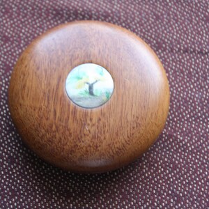 Op de afbeelding: Een ronde houten doos met een cirkelvormige inleg met een geschilderde boom met gele bloemen en groene bladeren. De inleg is omgeven door een zilveren rand.