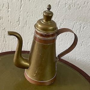 Puede incluir: Una cafetera vintage de latón y cobre con cuerpo redondeado y un pico largo. La cafetera tiene una banda decorativa alrededor del medio y una tapa con un pomo.
