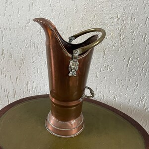 Könnte beinhalten: Ein kupferfarbener Kohleeimer mit Messinggriff und dekorativem Löwenkopfverschluss. Der Eimer hat einen ausgestellten Ausguss und einen abgerundeten Sockel mit silberfarbener Zierleiste. Es ist ein Vintage-Artikel, der wahrscheinlich zur Aufbewahrung von Kohle in der Nähe eines Kamins verwendet wurde.