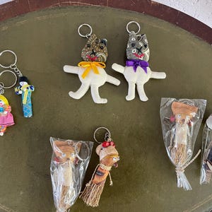 May include: A collection of handmade keychains featuring various designs. Includes fabric dolls with patterned heads and white bodies, and small figures in traditional clothing. Some are packaged in clear plastic.