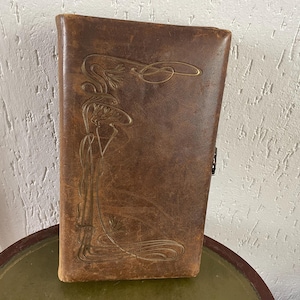 Puede incluir: Un libro antiguo encuadernado en cuero marrón con un diseño Art Nouveau en relieve en la portada. La portada presenta un diseño floral decorativo y líneas fluidas. El libro se cierra con un pequeño cierre metálico. El libro está sobre una mesa verde.