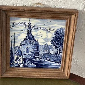 May include: A framed blue and white Delft tile depicting a tower with the text "HOORN HOOFDTOREN". The tile is surrounded by a wooden frame.