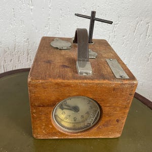 May include: A vintage wooden box with a clock face, metal handle, and a cross-shaped element on top. The box has a rectangular shape and a small metal plate. The clock face is visible through a circular opening.