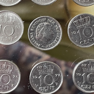 May include: A collection of Dutch 10-cent coins from the 1960s and 1970s. The coins are silver-colored and feature a crown, the number 10, and the word "CENT". Some coins have the year of minting visible, such as 1963, 1969, 1973, 1977, and 1979.