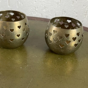 May include: Two antique brass candle holders. Each holder is a rounded shape with heart-shaped cutouts. The candle holders are on a green surface. The brass has a warm, aged patina.