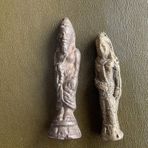 Op de afbeelding: Twee kleine, antieke beelden. Eén beeldt een figuur af met een hoge hoed en baard, de andere een vrouwelijke figuur met lang haar. Beide staan en hebben een verweerde, metalen uitstraling. De beelden staan op een getextureerd, olijfgroen oppervlak.