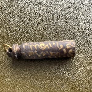 Puede incluir: Un colgante cilíndrico de color marrón oscuro con diseños grabados en color dorado. El colgante tiene un pequeño lazo en un extremo con un anillo dorado. El fondo es una superficie texturizada de color verde oliva.