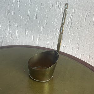 Puede incluir: Un cucharón de latón con un mango largo y adornado. El cucharón tiene forma ovalada y un acabado liso y pulido.