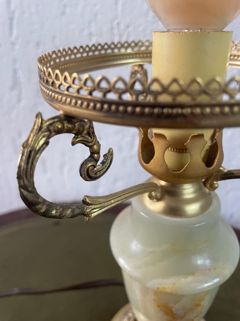 Op de afbeelding: Een vintage albasten tafellamp met een gouden metalen voet en sierlijke details. De lamp heeft een ronde, opengewerkte kaphouder en een decoratieve handgreep.