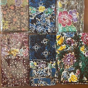 Puede incluir: Seis piezas de tela batik coloridas con estampados florales. Las telas presentan una variedad de colores, incluyendo rojo, azul, amarillo y verde, con diseños intrincados. Cada pieza está envuelta individualmente en plástico transparente.