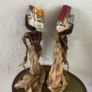 May include: Two Wayang Golek rod puppets featuring carved wooden heads with painted facial features, black hair, and elaborate headpieces. They wear purple bodices with gold sequins and long, flowing beige fabric skirts, each held by thin metal rods.