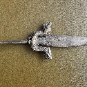 Puede incluir: Un objeto metálico oscuro con una estructura central ornamentada y una larga hoja puntiaguda. El objeto tiene una apariencia envejecida, lo que sugiere que es un artículo antiguo. Descansa sobre una superficie verde oliva texturizada.