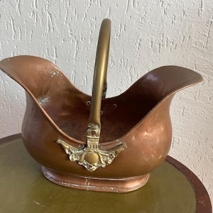 Op de afbeelding: Een koperen kolenkit met een messing handvat en decoratieve accenten. De kit heeft een brede, open bovenkant en een gebogen lichaam. De handgreep is bevestigd met sierlijke messing fittingen. Het stuk is waarschijnlijk antiek en vertoont tekenen van ouderdom.