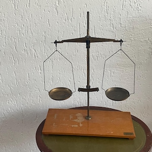 May include: An antique balance scale with a wooden base and brass components. The scale features two circular pans suspended by wire frames, connected to a central beam and vertical support. The scale is designed for measuring weight.