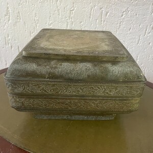 Puede incluir: Una caja cuadrada de latón antigua con intrincados diseños grabados. La caja tiene una tapa con bisagras y está decorada con un patrón floral.