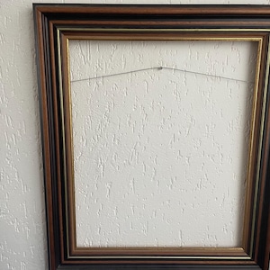 May include: A rectangular picture frame with a dark brown and black border, with a thin gold inner edge. A wire is strung across the back for hanging. The frame is empty, ready for a photograph or artwork.