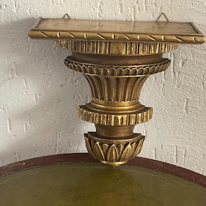 May include: A gold-colored, ornate wall shelf with a decorative, carved design. The shelf has a rectangular top and a detailed, column-like support structure. The shelf is mounted on a textured, white wall.