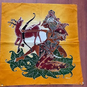 Puede incluir: Un tejido batik colorido que representa una criatura mítica con un arco y una flecha, cazando un ciervo. La criatura está rodeada de follaje verde y el fondo es de un amarillo vibrante.