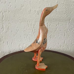 May include: Wooden duck figurine with a long neck and orange feet. The duck is made of light-colored wood with a patterned body and black accents. It stands on a green and brown table.