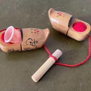 May include: Two wooden clogs with red and green painted flowers. One clog has a pink thimble inside and the other has a red felt ball. A red string connects the two clogs and a wooden stick.