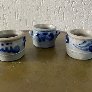 May include: Three small, blue and white ceramic crocks with handles. The crocks are decorated with blue and white floral patterns.