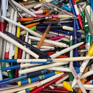 Può includere: Una grande collezione di penne colorate con vari loghi e testi. Le penne sono principalmente bianche, blu, rosse e gialle. Alcune delle scritte sulle penne includono "EXPO", "eurostar.com", "EUTERPE", "Chut Paradiso" e "Vedio!"