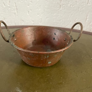 Könnte beinhalten: Eine kleine, antike Kupferschale mit zwei Drahtgriffen. Die Schale hat eine abgenutzte, gealterte Patina und ist leicht verbeult.