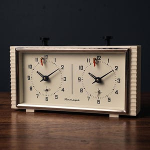 May include: A rectangular analog chess clock with a cream-colored casing and two circular clock faces. Each face features black numerals from 1 to 12, black hour and minute hands, and a small red indicator hand. The text "Янтарь" is printed in black.