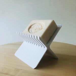 May include: A white, X-shaped soap dish holding a bar of soap. The soap is light beige with the text "HACISAKIR" embossed on it. The dish is made of a ridged plastic material.