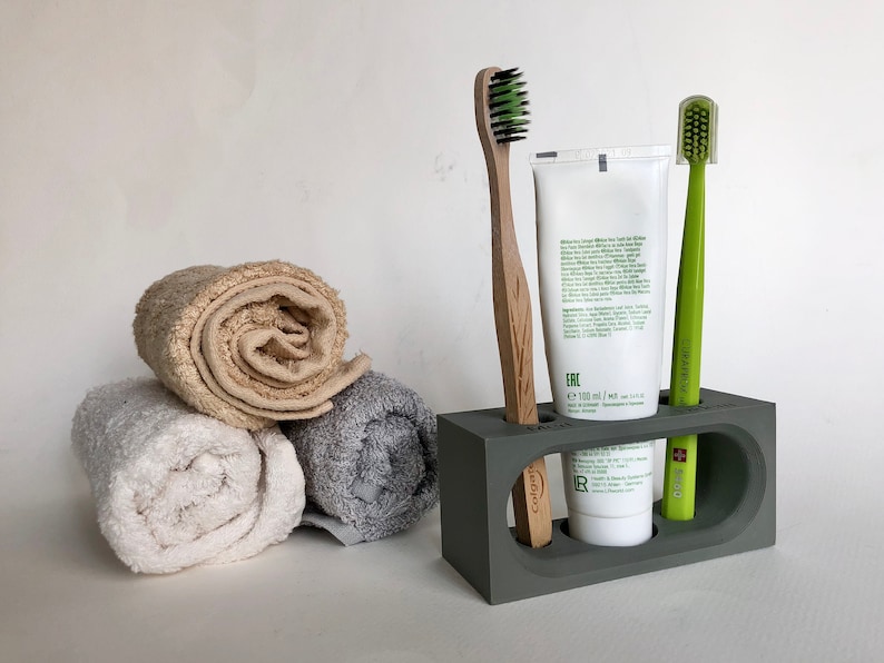 May include: A gray bathroom countertop organizer with three slots. The organizer holds a tube of toothpaste, a green toothbrush, and a wooden toothbrush. Two rolled towels are in the foreground.