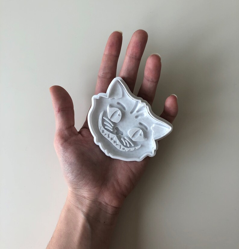 May include: A white plastic cookie cutter in the shape of the Cheshire Cat's face, featuring a wide grin and detailed facial features. The cutter is held in a hand against a plain background. This item is likely for baking.