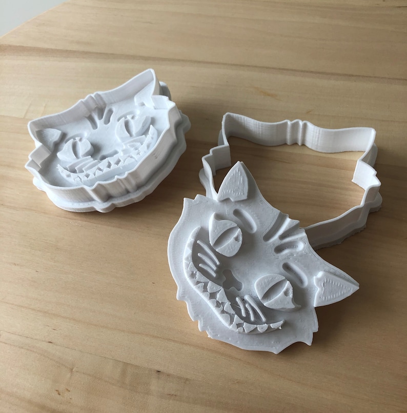 May include: Three white plastic cookie cutters in the shape of the Cheshire Cat's face from Alice in Wonderland. The cutters are on a light wood surface. One cutter is a complete face, and the other is the outline.
