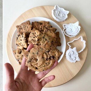 May include: A plate of cat-shaped cookies with chocolate chips and oats, alongside white plastic cookie cutters. One cookie is held in a hand. The cookies are on a wooden serving board, with a white table in the background.