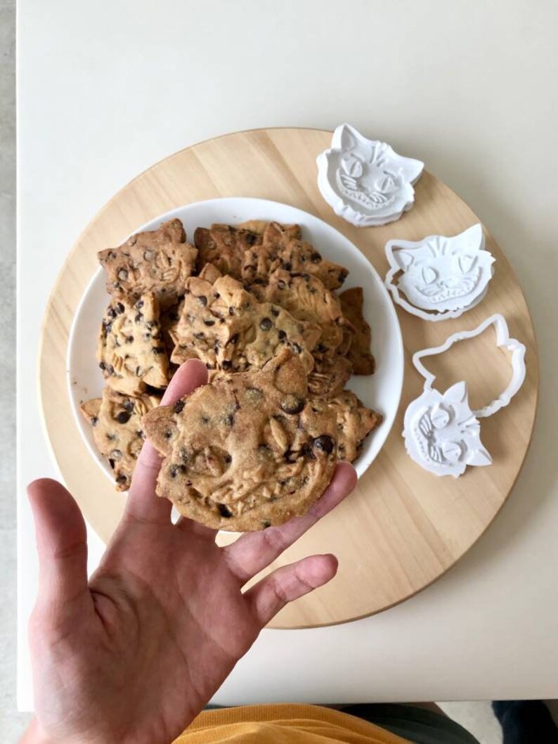 May include: A plate of cat-shaped cookies with chocolate chips and oats, alongside white plastic cookie cutters. One cookie is held in a hand. The cookies are on a wooden serving board, with a white table in the background.