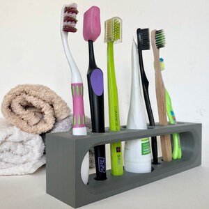May include: A gray toothbrush holder with six slots, holding a variety of toothbrushes, including a pink toothbrush with green accents, a black toothbrush with a purple accent, a green toothbrush with a white handle, a white toothbrush with a green handle, a black toothbrush with a white handle, and a wooden toothbrush with a green handle.