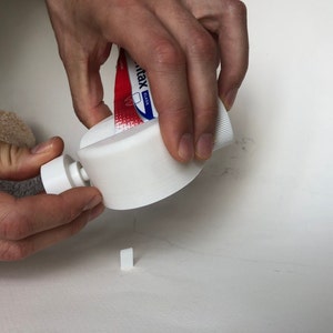 May include: A white 3D-printed toothpaste tube squeezer is being used to dispense toothpaste. The tube is red and white with blue text. A small, white, rectangular piece is on the surface.