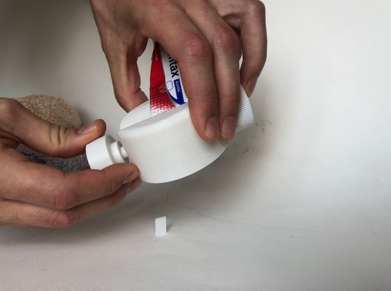 May include: A white 3D-printed toothpaste tube squeezer is being used to dispense toothpaste. The tube is red and white with blue text. A small, white, rectangular piece is on the surface.