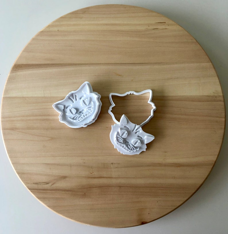 May include: Three white plastic cookie cutters in the shape of the Cheshire Cat's face, with a smiling expression. The cutters are on a light brown wooden surface. One cutter is a solid shape, and the other is an outline.