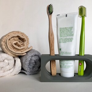 May include: A gray, rectangular toothbrush holder with two slots for toothbrushes and a space for a tube of toothpaste. The holder has a white tube of toothpaste with the brand name "Ere" and a volume of 100 ml. There are two toothbrushes, one with a green handle and the brand name "Curaprox" and the other with a wooden handle and the brand name "Colgate".