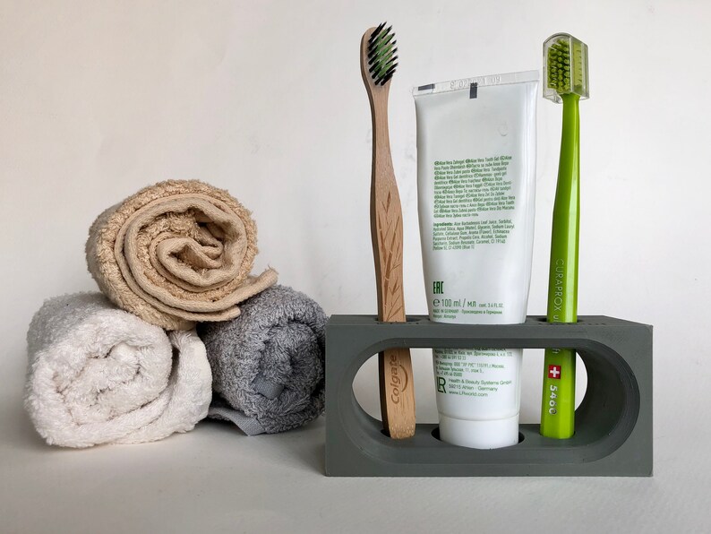 May include: A gray, rectangular toothbrush holder with two slots for toothbrushes and a space for a tube of toothpaste. The holder has a white tube of toothpaste with the brand name "Ere" and a volume of 100 ml. There are two toothbrushes, one with a green handle and the brand name "Curaprox" and the other with a wooden handle and the brand name "Colgate".