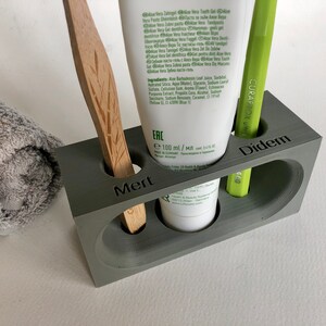 May include: A gray, rectangular toothbrush holder with three compartments. The holder has the names "Mert", "Didem", and "ERC" printed on the front. The holder contains a wooden toothbrush, a green toothbrush, and a tube of toothpaste.
