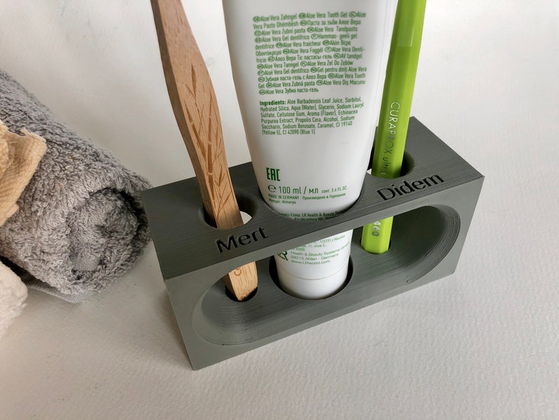 May include: A gray, rectangular toothbrush holder with three compartments. The holder has the names "Mert", "Didem", and "ERC" printed on the front. The holder contains a wooden toothbrush, a green toothbrush, and a tube of toothpaste.