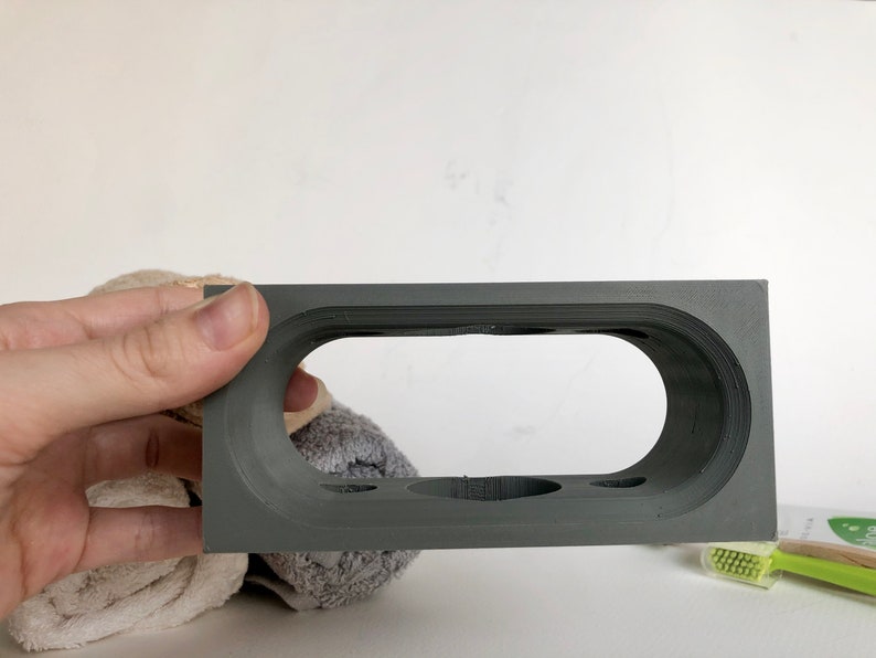 May include: Gray 3D printed toothbrush holder with four slots and a large oval opening. The holder is made of a smooth, matte material.