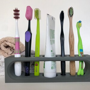 May include: A gray toothbrush holder with seven slots, holding six toothbrushes and a tube of toothpaste. The toothbrushes are a variety of colors and styles, including a pink toothbrush with green accents, a black toothbrush with a purple handle, a green toothbrush with a white handle, a black toothbrush with a black handle, a wooden toothbrush with a brown handle, and a green toothbrush with a blue handle. The toothpaste tube is white with black text.
