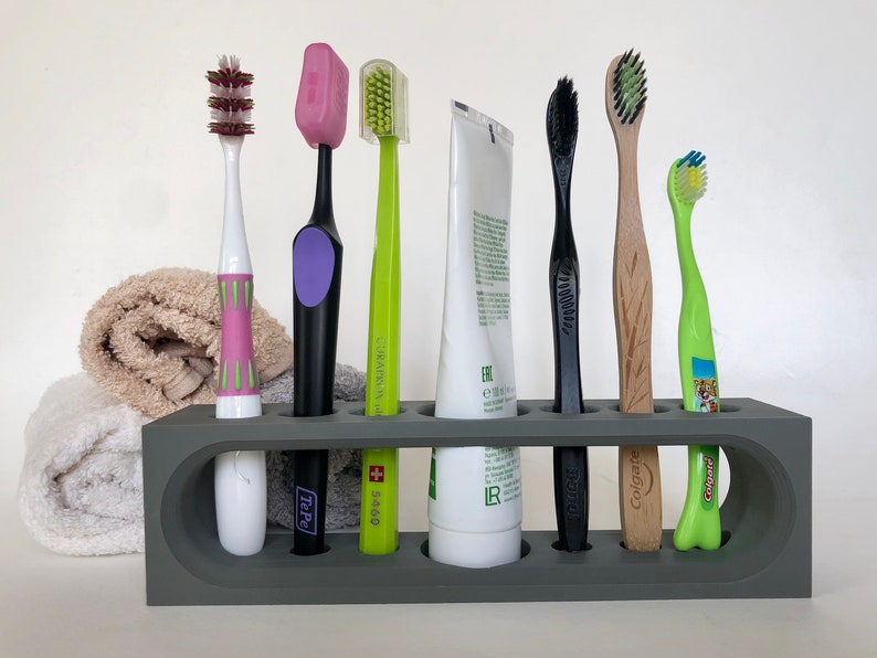 May include: A gray toothbrush holder with seven slots, holding six toothbrushes and a tube of toothpaste. The toothbrushes are a variety of colors and styles, including a pink toothbrush with green accents, a black toothbrush with a purple handle, a green toothbrush with a white handle, a black toothbrush with a black handle, a wooden toothbrush with a brown handle, and a green toothbrush with a blue handle. The toothpaste tube is white with black text.