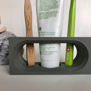 May include: Gray bathroom countertop organizer with three slots. A white tube of toothpaste with green lettering that says "ERC e 100 ml/Ml" is in the center slot. A green toothbrush with white lettering that says "CURAPROX +5460" is in the right slot. A wooden toothbrush with green lettering that says "Colgate" is in the left slot.